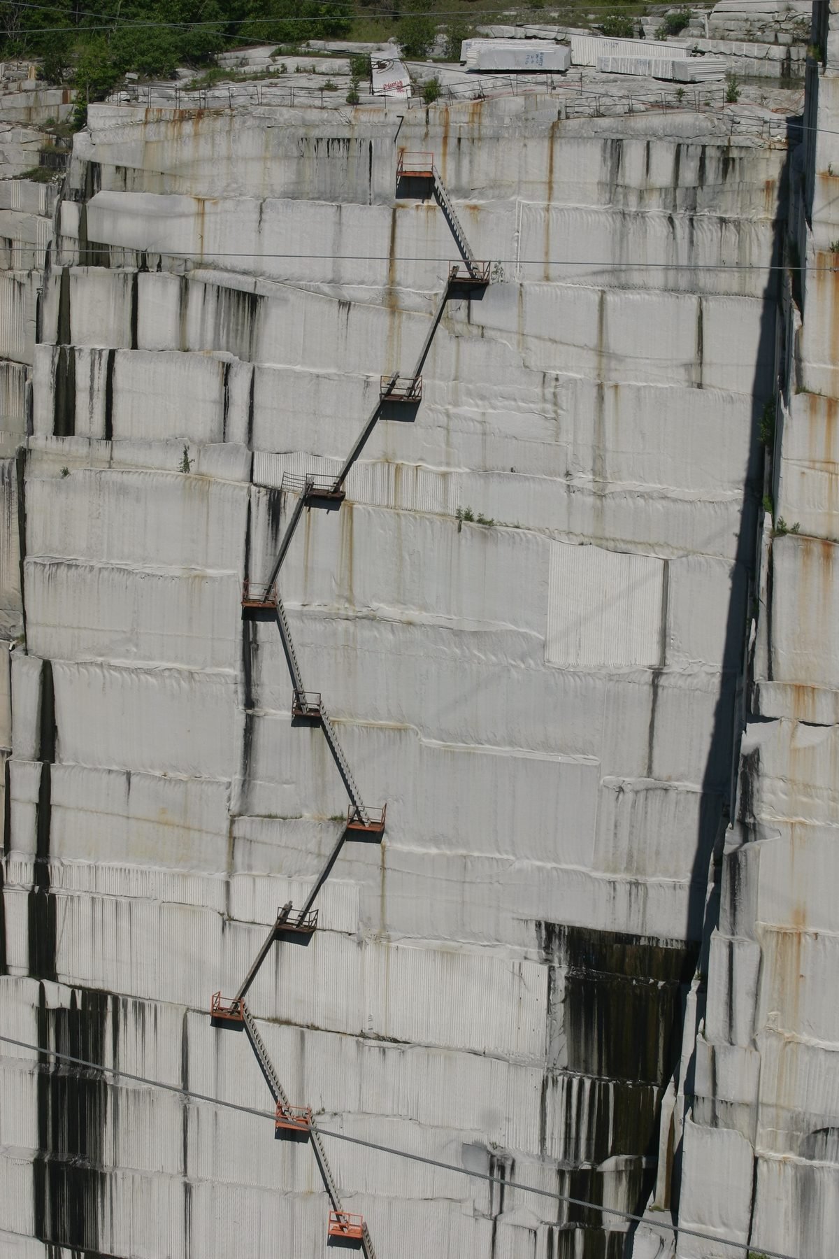 A pale granite quarry wall cut in tall benches, with a zigzag of ladders down its face